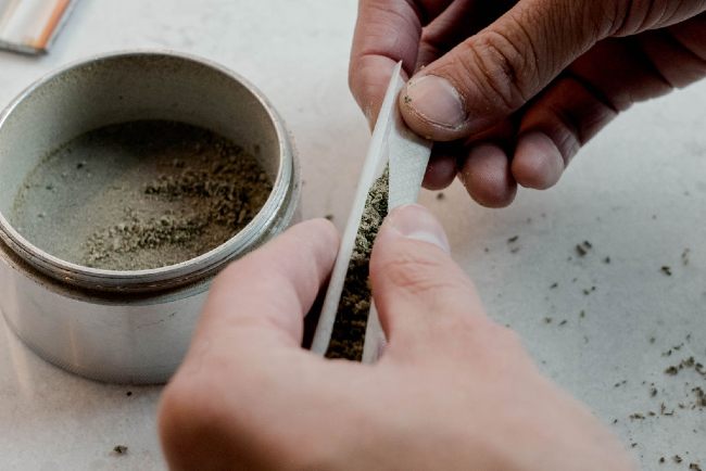 a person’s hands about to roll a joint that has been stuffed with ground cannabis, next to an almost empty silver grinder